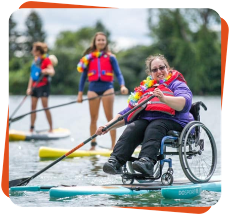 Trois femmes font de la planche à pagaie. Celles en arrière-plan sont debout sur leur planche régulière. La femme en avant-plan est assise dans un fauteuil roulant et le fauteuil est sur une planche à pagaie adaptée.